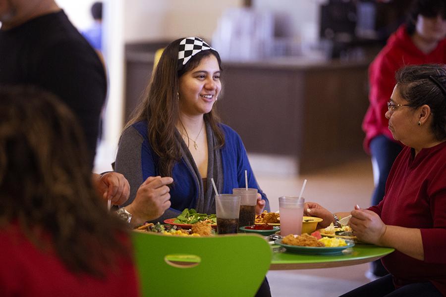 A group of people in a dining hall sit at a table with lime green chairs and several plates of food on the table.